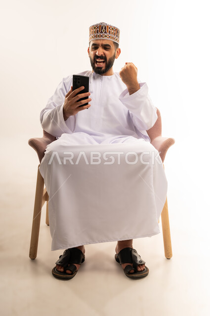 رفع قبضة اليد للاعلى بتعابير الفوز والنصر، إيماءات السعادة والحماس بالنجاح وتحقيق الهدف، مفهوم تلقي الخبر الجيد, رجل عربي خليجي عماني يرتدي الدشداشة والكمة يجلس على كرسى مريح ، يحمل بيده الهاتف المحمول، إنجاز المهام المطلوبة, استخدام جهاز تقني حديث ومتطور ، خلفية بيضاء.