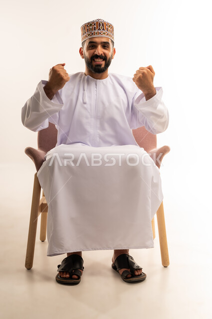 رفع قبضة اليد بتعابير الفوز والانتصار، الحماس لتحقيق الاهداف ، بورتريه لشاب عربي خليجي عماني يرتدي الدشداشة والكمة يجلس على كرسى مريح، ينظر إلى الكاميرا بايماءات الفرح والسرور، خلفية بيضاء .