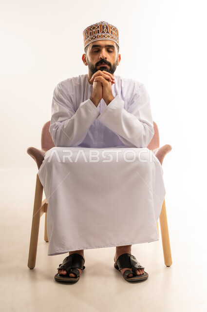 مفهوم الاسترخاء وشرود الذهن و التأمل , النعاس والحاجة الى النوم والراحة ،  إيجاد حل لمشكلة ما, بورتريه لرجل عربي خليجي عماني يرتدي الكمة والدشداشة يجلس على كرسي مريح يضع يده على ذقنه بايماءات الجدية، خلفية بيضاء .