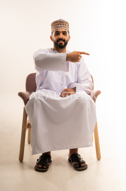 Defining and clarifying a specific location, pointing to an item, portrait of a smiling young Omani Arab man wearing a keffiyeh and a dishdasha, sitting on a comfortable chair, pointing with his index finger to a specific target, white background.