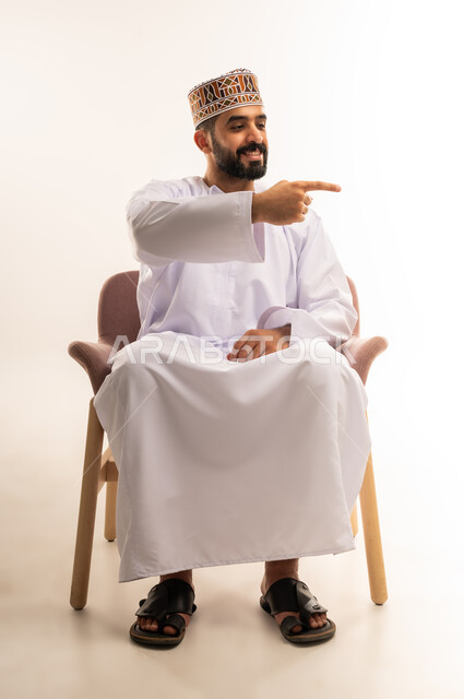 النظر إلى شيء ما بتعابير السرور, الإشارة إلى هدف محدد, بورتريه لشاب عربي خليجي عماني مبتسم يرتدي الكمه و الدشداشة يجلس على كرسى مريح ، يرفع اصبع السبابة لتحديد وتوضيح موقع معين, خلفية بيضاء .