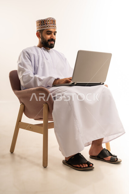 عقد إجتماع عمل عبر اللابتوب ، استعمال جهاز اللابتوب والتقنيات الحديثة فى انجاز المهام ،دمج التقنية والتكنولوجيا المتطورة بالعمل، مهنة ووظيفة مكتبية، بورتريه لرجل عربي خليجي عماني يرتدي الدشداشة والكمه يجلس على كرسي مريح يعمل على جهاز الحاسوب المحمول، خلفية بيضاء .