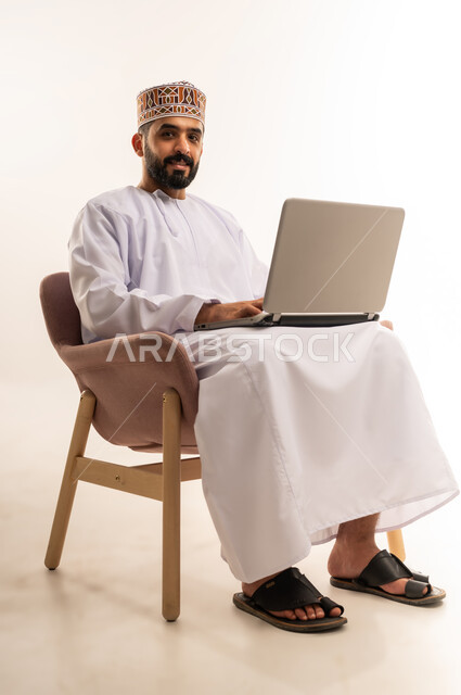 انجاز مهام العمل عن بعد ، استعمال جهاز اللابتوب والتقنيات الحديثة ،دمج التقنية والتكنولوجيا المتطورة بالعمل، مهنة ووظيفة مكتبية، بورتريه لرجل عربي خليجي عماني يرتدي الدشداشة والكمه يجلس على كرسي مريح يعمل على جهاز الحاسوب المحمول، خلفية بيضاء .