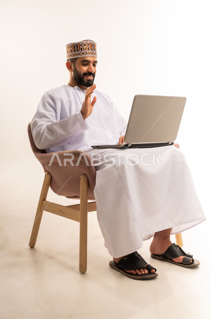 القاء التحية عبر مكالمات الفيديو ، دمج التقنية والتكنولوجيا المتطورة بالعمل، استعمال جهاز اللابتوب والتقنيات الحديثة في إنجاز المهام ، مهنة ووظيفة مكتبية، بورتريه لرجل عربي خليجي عماني يرتدي الدشداشة والكمه يجلس على كرسي مريح يعمل على جهاز الحاسوب المحمول ، خلفية بيضاء .