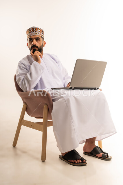 دمج التقنية والتكنولوجيا المتطورة بالعمل، استعمال جهاز اللابتوب والتقنيات الحديثة في إنجاز المهام ، مهنة ووظيفة مكتبية، بورتريه لرجل عربي خليجي عماني يرتدي الدشداشة والكمه يجلس على كرسي مريح يعمل على جهاز الحاسوب المحمول ، خلفية بيضاء .