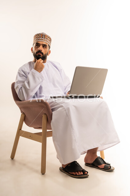 استعمال جهاز اللابتوب والتقنيات الحديثة في إنجاز المهام ، مهنة ووظيفة مكتبية، دمج التقنية والتكنولوجيا المتطورة بالعمل، بورتريه لرجل عربي خليجي عماني يرتدي الدشداشة والكمه يجلس على كرسي مريح يعمل على جهاز الحاسوب المحمول ، خلفية بيضاء .