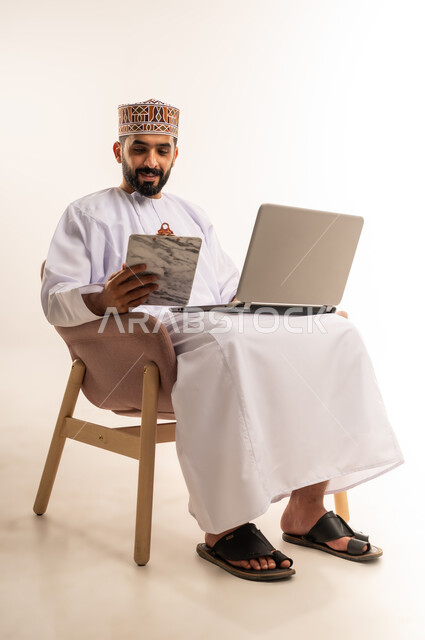 مهنة ووظيفة مكتبية، دمج التقنية والتكنولوجيا المتطورة بالعمل، بورتريه من الخلف لرجل عربي خليجي عماني يرتدي الدشداشة والكمه يجلس على كرسي مريح يعمل على جهاز الحاسوب المحمول ويحمل بيده الاوراق ، خلفية بيضاء .