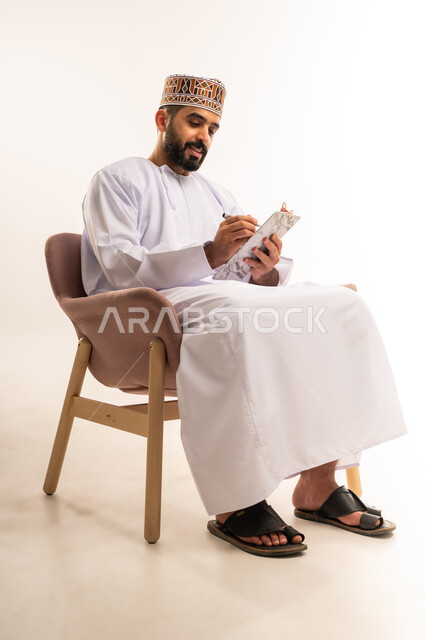 Using paper to record important notes and information, tools for writing down daily tasks, portrait of a young Omani Gulf Arab man wearing a dishdasha and a kuma, holding a piece of paper and a pen in his hand, writing something with gestures of concentration and contemplation, white background.