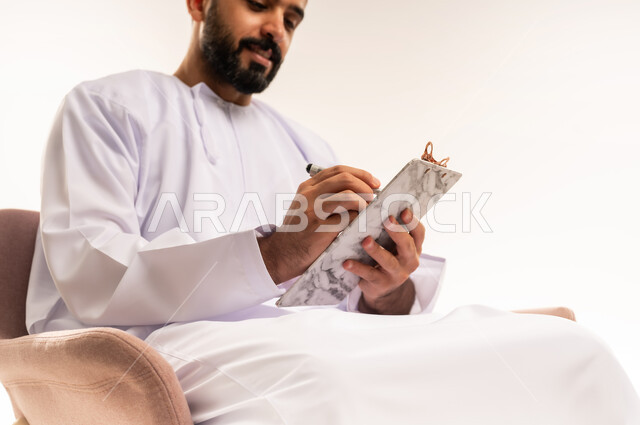 Daily task writing tools, using paper to record notes and important information, close-up portrait of a young Omani Gulf Arab man wearing a dishdasha and a kuma holding a piece of paper and a pen, writing something with gestures of concentration and contemplation, white background.