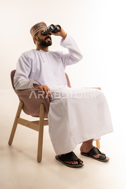 Love of exploration and adventure, using binoculars to discover distant objects and bring them closer, portrait of a young Omani Gulf Arab man wearing a kuma and a dishdasha sitting on a comfortable chair holding binoculars and looking at something, gestures of enjoyment and pleasure, white background.