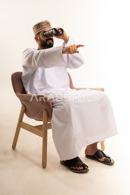 استعمال المنظار للكشف عن الاشياء البعيدة وتقريبها, بورتريه لشاب عربي خليجي عماني يرتدي الكمه والدشداشة يجلس على كرسي مريح يحمل بيده المنظار ثنائي العينين وينظر الى شيء ما، حب الاستكشاف والمغامرة، إيماءات الاستمتاع والسرور، خلفية بيضاء .