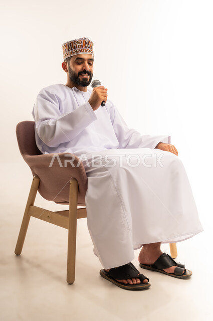ممارسة المواهب الفنية ، ايماءات وجه و يدين تدل على السعادة و الاستمتاع ، بورتريه لشاب عربي خليجي عماني يرتدي الدشداشة والكمة ، يجلس على كرسى ويحمل الميكروفون ويقوم بالغناء بحماس ، خلفية بيضاء .