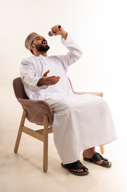 ايماءات وجه و يدين تدل على السعادة و الاستمتاع ، بورتريه لشاب عربي خليجي عماني يرتدي الدشداشة والكمة ، يجلس على كرسى ويحمل الميكروفون ويقوم بالغناء بحماس ، خلفية بيضاء .