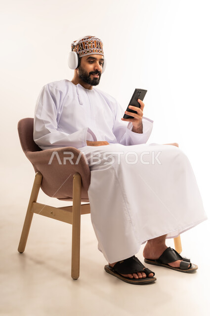 استخدام الهاتف و تقنية الهيدفون ، الاستماع إلى الموسيقى والتسجيلات ، استخدام البرامج الصوتية ، الاستفادة من الجوال والأجهزة التكنولوجية، بورتريه لشاب عربي خليجي عماني يرتدي الدشداشة والكمة ، يجلس على كرسى ويضع سماعة الرأس بإيماءات السعادة، خلفية بيضاء .