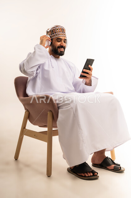 استخدام البرامج الصوتية  ، الاستماع إلى الموسيقى والتسجيلات ، الاستفادة من الجوال والأجهزة التكنولوجية، بورتريه لشاب عربي خليجي عماني يرتدي الدشداشة والكمة ، يجلس على كرسى ويمسك سماعة الرأس بإيماءات السعادة, استخدام الهاتف و تقنية الهيدفون، خلفية بيضاء .