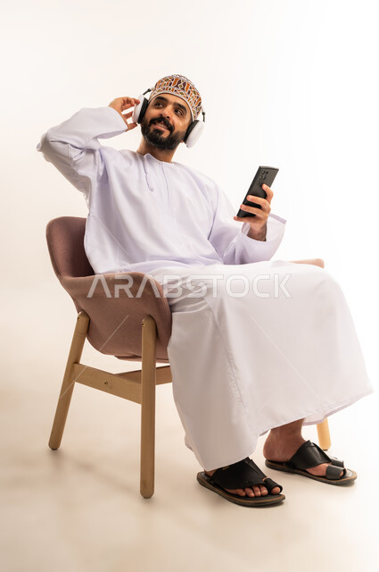 الاستماع إلى الموسيقى والتسجيلات الصوتية، الاستفادة من الجوال والأجهزة التكنولوجية، بورتريه لشاب عربي خليجي عماني يرتدي الدشداشة والكمة ، يجلس على كرسى ويمسك سماعة الرأس بإيماءات السعادة, استخدام الهاتف و تقنية الهيدفون، خلفية بيضاء .