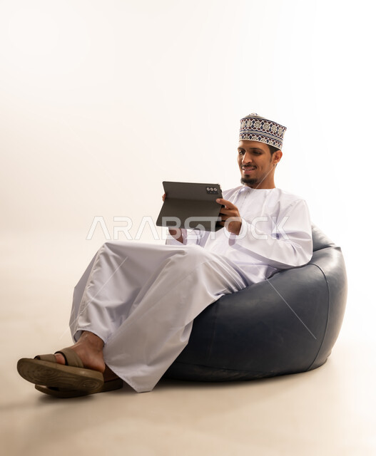 Enjoying reading, portrait of a young Omani Gulf Arab man wearing a dishdasha and an Omani kummah sitting on a comfortable chair reading stories and browsing scientific books, white background