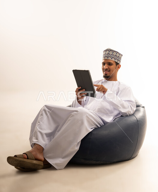 Technological development in various fields, the use of modern technology in remote work, holding meetings via the Internet, completing work quickly and easily, a portrait of an Arab Gulf Omani man wearing a cufflink and a dishdasha holding a tablet in his hand, white background