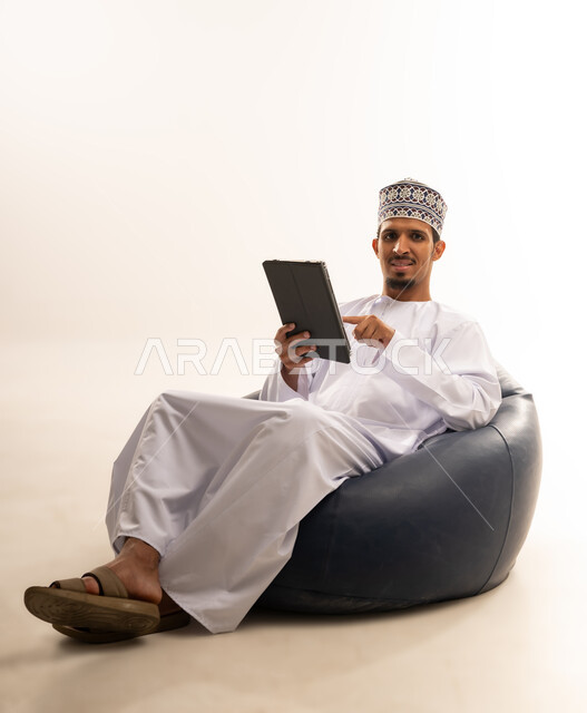 التطور التقني في مختلف المجالات, استخدام التكنولوجيا الحديثة في العمل عن بعد، عقد الاجتماعات عن طريق الانترنت، إنجاز الأعمال بسرعة وسهولة، بورتريه  لرجل عربي خليجي عماني يرتدي الكمه و الدشداشة يحمل بيده جهاز التابليت, خلفيه بيضاء