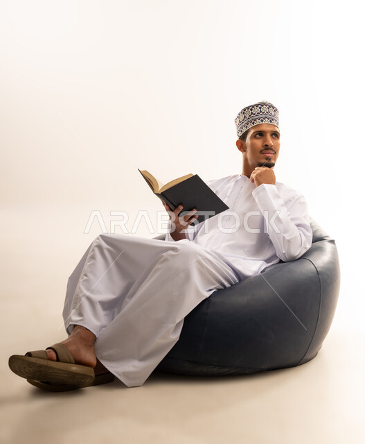 الاستمتاع بممارسة هواية المطالعة، بورتريه لشاب عربي خليجي عماني يرتدي الدشداشة والكمه العمانية يجلس على كرسي مريح يقوم بقراءة القصص وتصفح الكتب العلميه, خلفية بيضاء