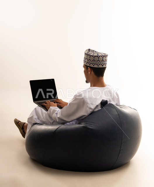 الاستمتاع بممارسة هواية المطالعة، استخدام الاجهزه التقنيه الحديثه في التعلم عن بعد, بورتريه من الخلف لشاب عربي خليجي عماني يرتدي الدشداشة والكمه العمانية يجلس على كرسي مريح يقوم بتصفح الابتوب, خلفية بيضاء