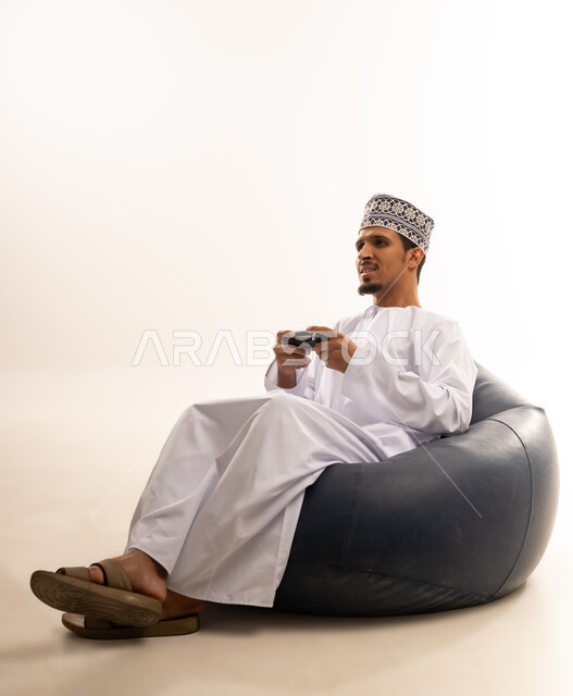 Having a fun and entertaining time, interacting and merging with PlayStation games, portrait of a young Saudi Omani man wearing a dishdasha and a kurta sitting on a comfortable chair and holding a joystick in his hand, white background