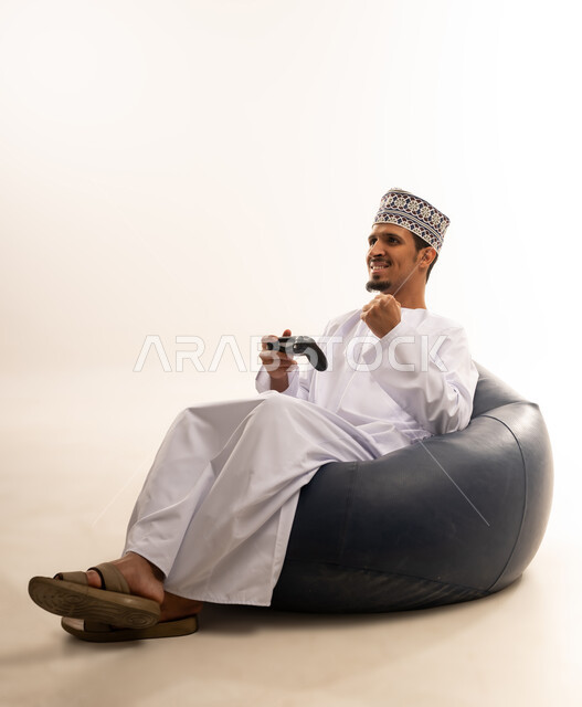 Having a fun and entertaining time, interacting and merging with PlayStation games, portrait of a young Saudi Omani man wearing a dishdasha and a kurta sitting on a comfortable chair and holding a joystick in his hand, white background