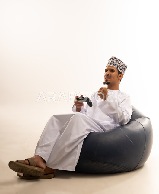 Having a fun and entertaining time, interacting and merging with PlayStation games, portrait of a young Saudi Omani man wearing a dishdasha and a kurta sitting on a comfortable chair and holding a joystick in his hand, white background