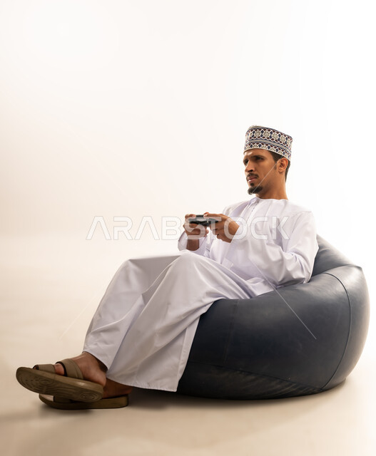 Having a fun and entertaining time, interacting and merging with PlayStation games, portrait of a young Saudi Omani man wearing a dishdasha and a kurta sitting on a comfortable chair and holding a joystick in his hand, white background