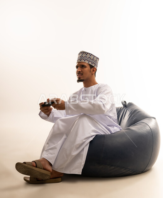 Having a fun and entertaining time, interacting and merging with PlayStation games, portrait of a young Saudi Omani man wearing a dishdasha and a kurta sitting on a comfortable chair and holding a joystick in his hand, white background