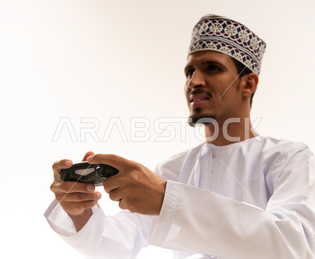 قضاء اوقات ممتعه ومسليه، التفاعل والاندماج مع ألعاب البلايستيشن، بورتريه مقرب لشاب عربي سعودي عماني يرتدي الدشداشه والكمه يحمل بيده عصا التحكم, خلفيه بيضاء