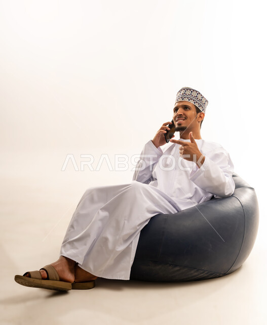استخدام الأجهزة والتقنيات الحديثة، الدردشة والتواصل مع الاصدقاء بإيماءات السعادة والفرح، بورتريه لرجل عربي خليجي عماني يرتدي الدشداشة والكمه يمسك بيده الهاتف المحمول يقوم بإجراء المكالمات الهاتفية، خلفية بيضاء