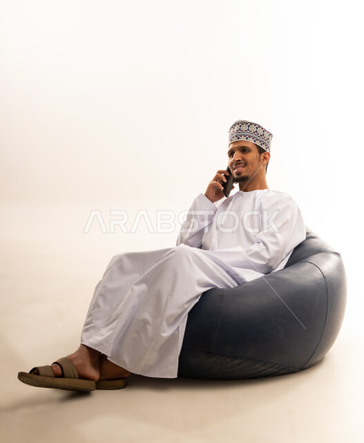 استخدام الأجهزة والتقنيات الحديثة، الدردشة والتواصل مع الاصدقاء بإيماءات السعادة والفرح، بورتريه لرجل عربي خليجي عماني يرتدي الدشداشة والكمه يمسك بيده الهاتف المحمول يقوم بإجراء المكالمات الهاتفية، خلفية بيضاء