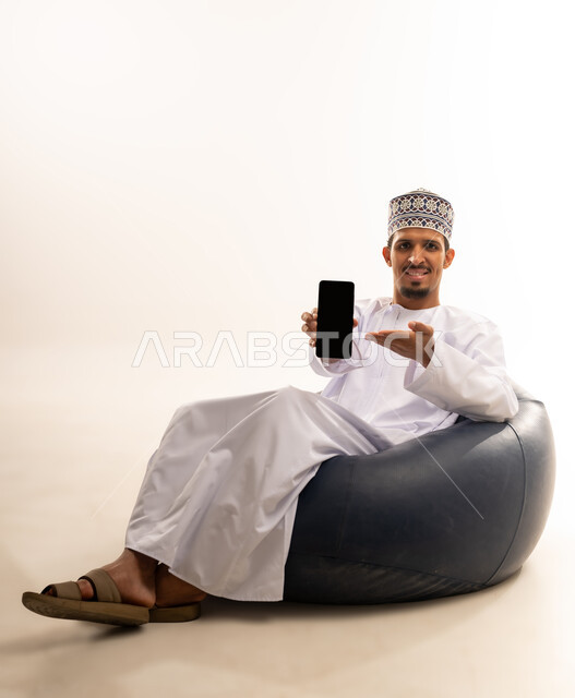 Mobile applications and services presentation, digital and electronic marketing concept, portrait of an Arab Gulf Omani man wearing a dishdasha and a kuma sitting on a luxurious leather sofa pointing with his mobile phone with a blank black screen, use of advanced technology, white background