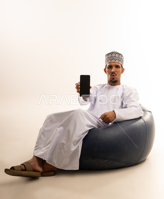 Mobile applications and services presentation, digital and electronic marketing concept, portrait of an Arab Gulf Omani man wearing a dishdasha and a kuma sitting on a luxurious leather sofa pointing with his mobile phone with a blank black screen, use of advanced technology, white background