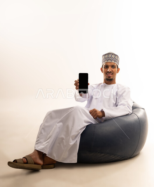 Mobile applications and services presentation, digital and electronic marketing concept, portrait of an Arab Gulf Omani man wearing a dishdasha and a kuma sitting on a luxurious leather sofa pointing with his mobile phone with a blank black screen, use of advanced technology, white background