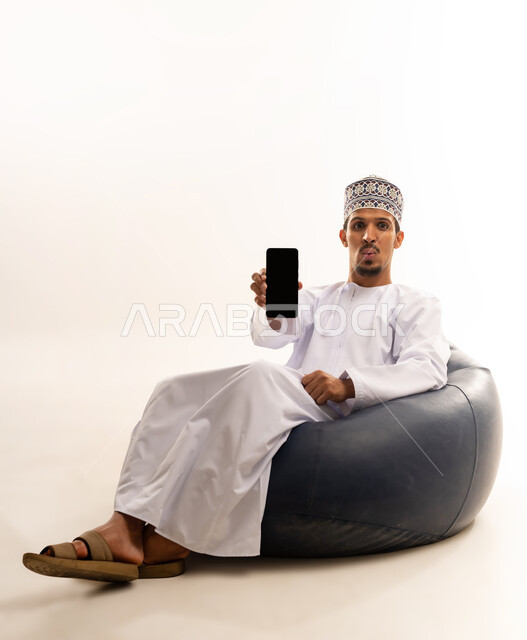 عرض تطبيقات وخدمات الهاتف المحمول، مفهوم التسويق الرقمي و الالكتروني، بورتريه لرجل عربي خليجي عماني يرتدي الدشداشة والكمة يجلس على اريكه من الجلد الفاخر يشير بجواله بشاشة سوداء فارغة، استخدام التكنولوجيا المتطورة، خلفية بيضاء