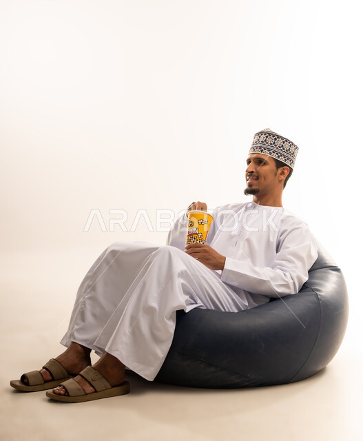 الاستمتاع بتناول السناك خلال أوقات الفراغ، بورتريه لشاب عربي خليجي عماني يرتدي الدشداشه والكمه يجلس على اريكه من الجلد ويقوم بتناول الفشار بايماءات الدهشه والسرور،  التسلية والاستمتاع بقضاء الاوقات الممتعه بمشاهده التلفاز، خلفيه بيضاء