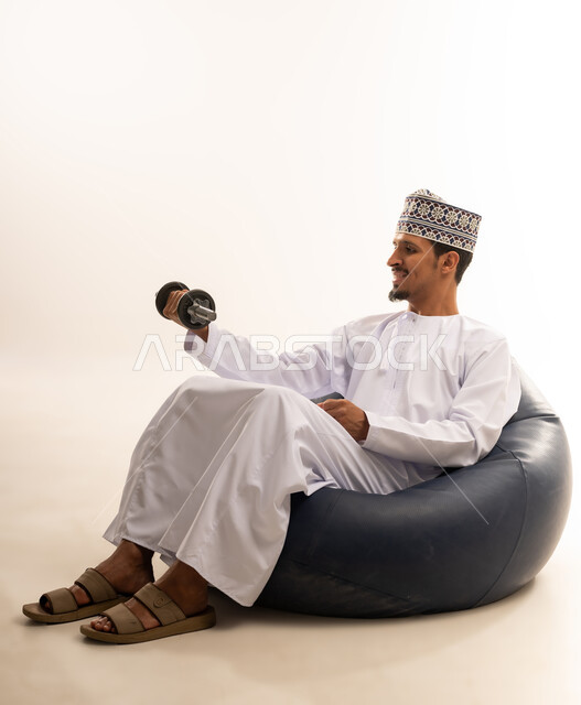رفع الأوزان الحديدية الخفيفه، أداء التمارين الرياضيه والانشطه الترفيهيه, بورتريه لرجل عربي خليجي عماني يرتدي الدشداشة والكمه يجلس على اريكه مريحه يحمل بيده الأثقال، نشاط بدني للمحافظة على الصحة و اللياقة الجسدية، بناء عضلات الجسم وتقويتها، خلفية بيضاء