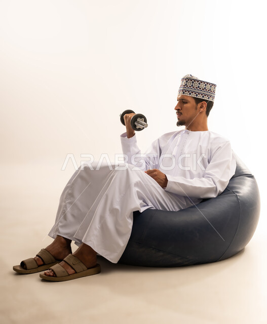 Lifting light iron weights, performing sports exercises and recreational activities, portrait of an Arab Gulf Omani man wearing a dishdasha and a cloak sitting on a comfortable sofa holding weights in his hand, physical activity to maintain health and physical fitness, building and strengthening the body muscles, white background