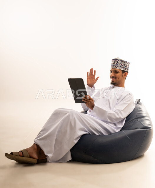 التواصل و الدردشة مع الاقارب, القيام بمقابلات وإجتماعات العمل أونلاين، استخدام جهاز تقني حديث لتصفح الصور والفيديوهات بسهولة، بورتريه  لرجل عربي خليجي عماني يرتدي الدشداشه والكمه يمسك بيده جهاز التابلت بشاشه سوداء فارغة, خلفية بيضاء