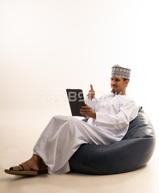 التواصل و الدردشة مع الاقارب, القيام بمقابلات وإجتماعات العمل أونلاين، استخدام جهاز تقني حديث لتصفح الصور والفيديوهات بسهولة، بورتريه  لرجل عربي خليجي عماني يرتدي الدشداشه والكمه يمسك بيده جهاز التابلت بشاشه سوداء فارغة, خلفية بيضاء