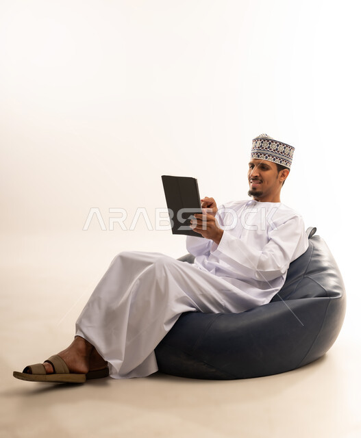 التواصل و الدردشة مع الاقارب, القيام بمقابلات وإجتماعات العمل أونلاين، استخدام جهاز تقني حديث لتصفح الصور والفيديوهات بسهولة، بورتريه  لرجل عربي خليجي عماني يرتدي الدشداشه والكمه يمسك بيده جهاز التابلت بشاشه سوداء فارغة, خلفية بيضاء