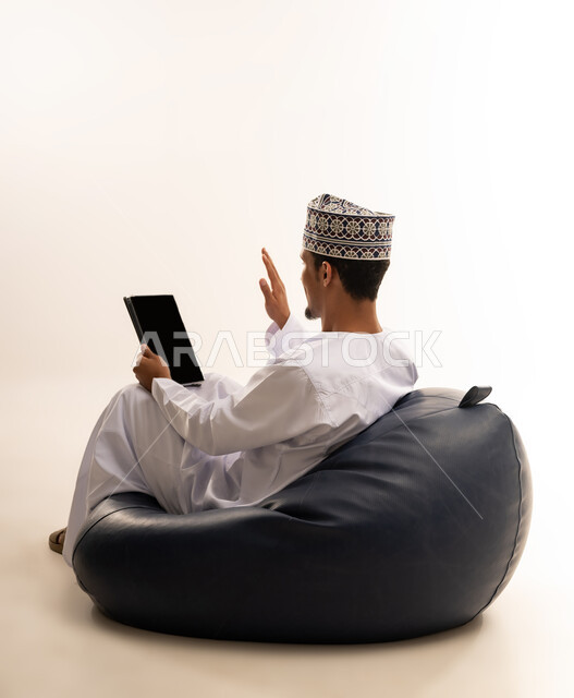 التواصل و الدردشة مع الاقارب, القيام بمقابلات وإجتماعات العمل أونلاين، استخدام جهاز تقني حديث لتصفح الصور والفيديوهات بسهولة، بورتريه مقرب من الخلف لرجل عربي خليجي عماني يرتدي الدشداشه والكمه يمسك بيده جهاز التابلت بشاشه سوداء فارغة, خلفية بيضاء