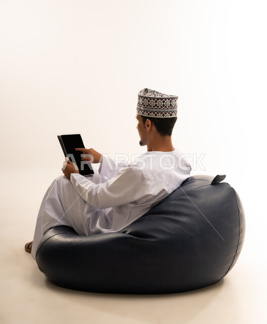 Communicating and chatting with relatives, conducting online interviews and business meetings, using a modern technical device to browse photos and videos easily, a close-up portrait from the back of an Arab Gulf Omani man wearing a dishdasha and a turban holding a tablet with a blank black screen, white background