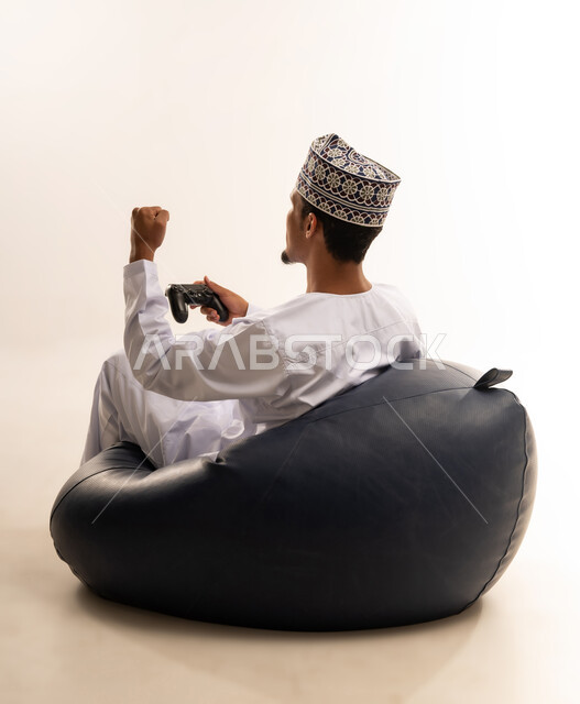 رفع قبضة اليد للاعلى بتعابير الفوز و الانتصار, قضاء وقت ممتع ومسلي، التفاعل والاندماج مع ألعاب الفيديو والبلايستيشن، بورتريه من الخلف لشاب عربي عماني خليجي يرتدي الكمة و الدشداشة يجلس على كرسي مريح ويحمل بيده عصا التحكم، خلفية بيضاء