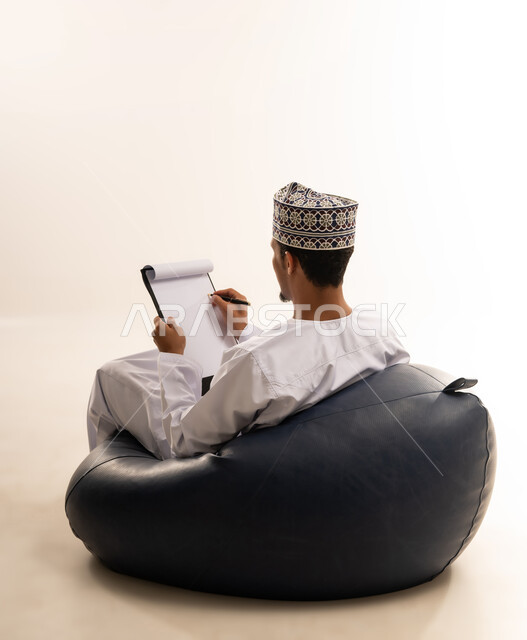 Expressions of thinking for decision making, taking notes, portrait of an Omani Arab Gulf man wearing a dishdasha and a kuma holding a notebook and pen in his hands writing information with gestures of optimism and happiness, white background