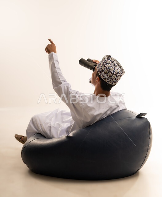 حب الاستكشاف و المغامرة, رؤية الاشياء البعيدة و تقريبها, بورتريه لشاب عربي خليجي عماني مبتسم يرتدي الدشداشة و الكمه يجلس على اريكه مريحه من الجلد الفاخر يحمل بيده منظار ثنائي العينين و ينظر الى شيء ما بإيماءات السرور والاستمتاع، خلفية بيضاء