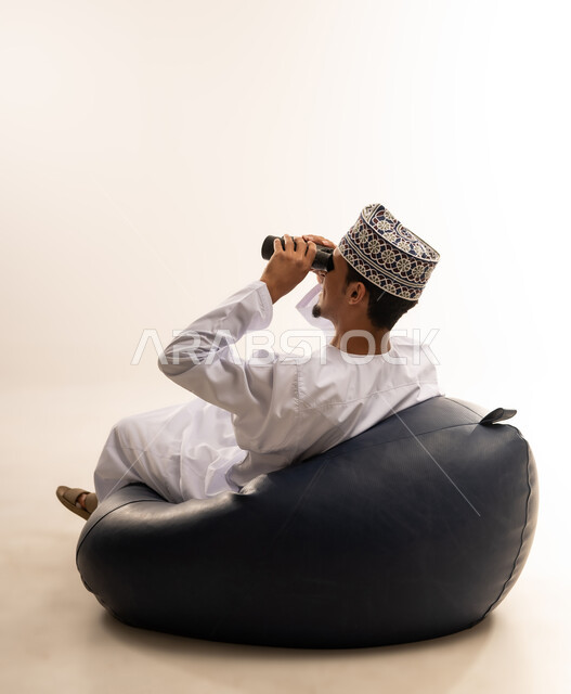 حب الاستكشاف و المغامرة, رؤية الاشياء البعيدة و تقريبها, بورتريه لشاب عربي خليجي عماني مبتسم يرتدي الدشداشة و الكمه يجلس على اريكه مريحه من الجلد الفاخر يحمل بيده منظار ثنائي العينين و ينظر الى شيء ما بإيماءات السرور والاستمتاع، خلفية بيضاء