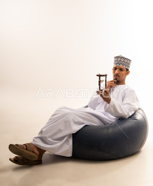 القيام بمراقبة توقيت الرمل, إدراك قيمة اللحظات الثمينة, مفهوم إدارة الأعمال وتنظيم الجدول الزمني, بورتريه لشاب عربي خليجي عماني يرتدي الكمة و الدشداشة يجلس على كرسي مريح يراقب الساعة الرملية الزجاجية (البنكام) وينظر إليها, خلفية بيضاء